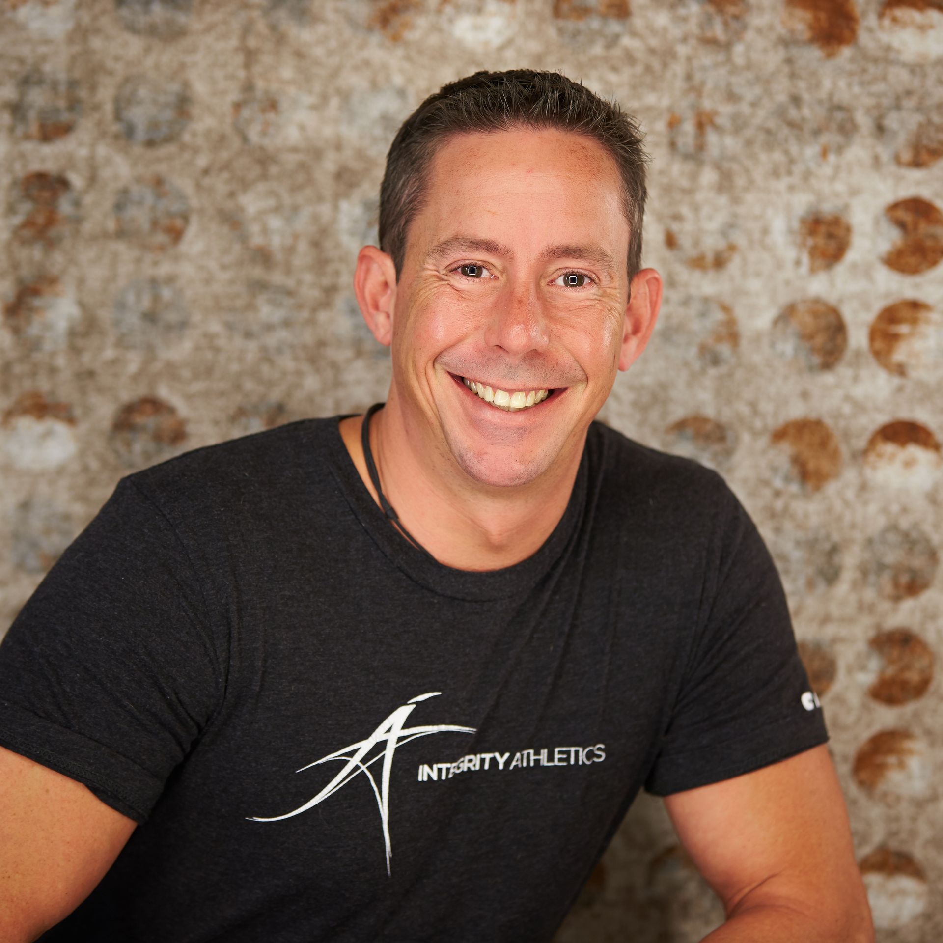 Smiling person in a black T-shirt against a textured stone wall.