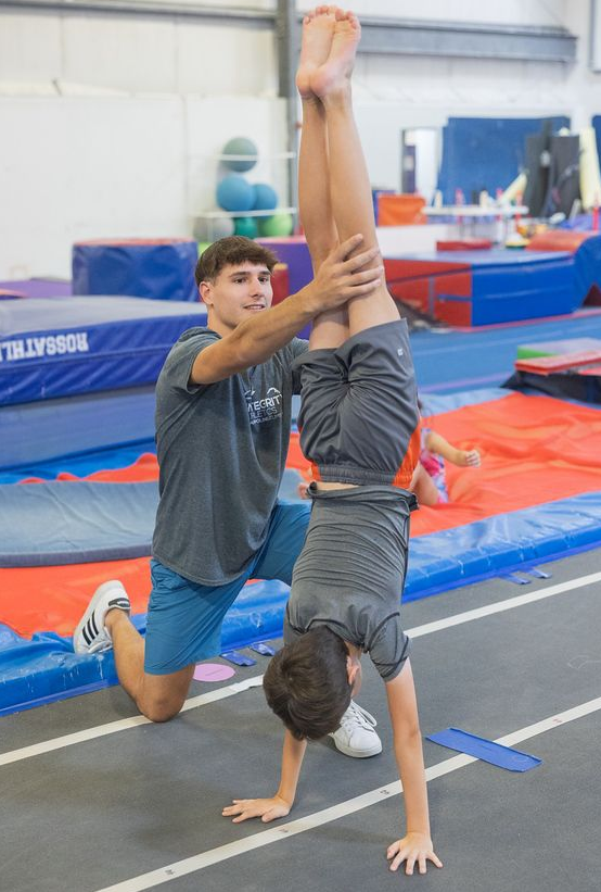 A man is helping a young girl do a handstand on a blue mat