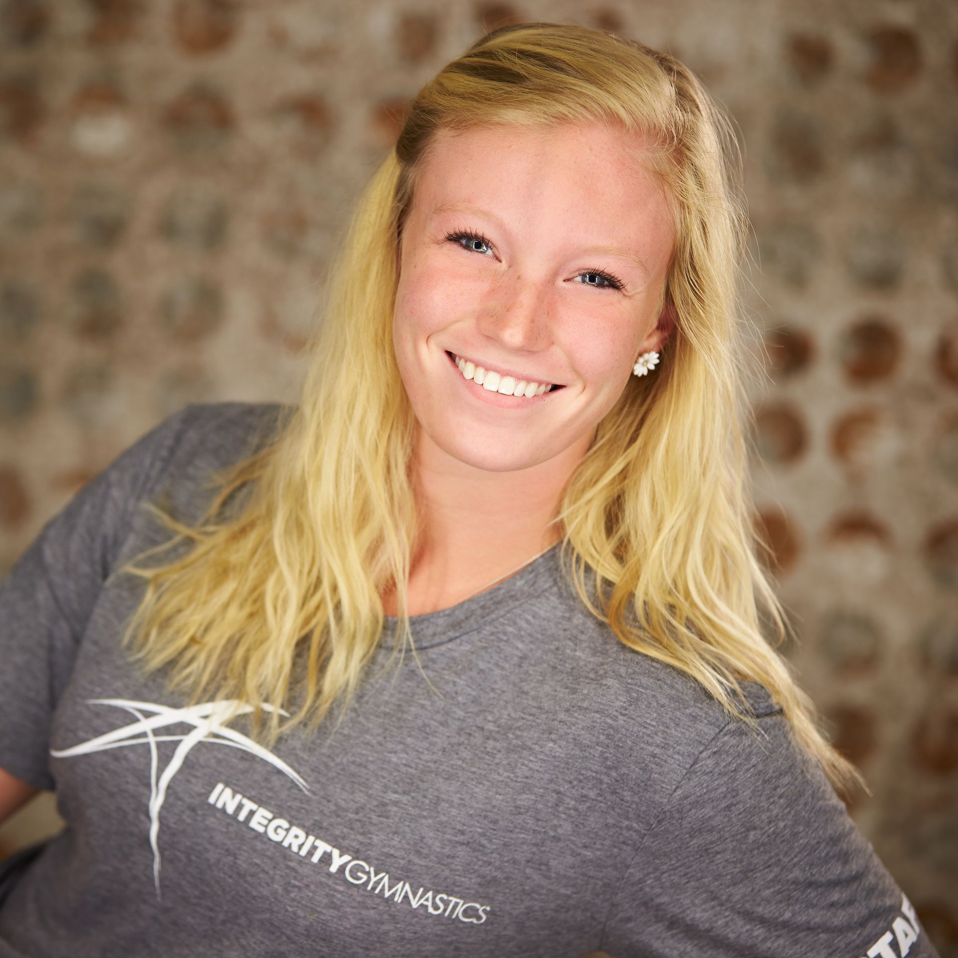 Smiling blonde athlete in a gray Integrity Gymnastics T-shirt against a blurred brick background