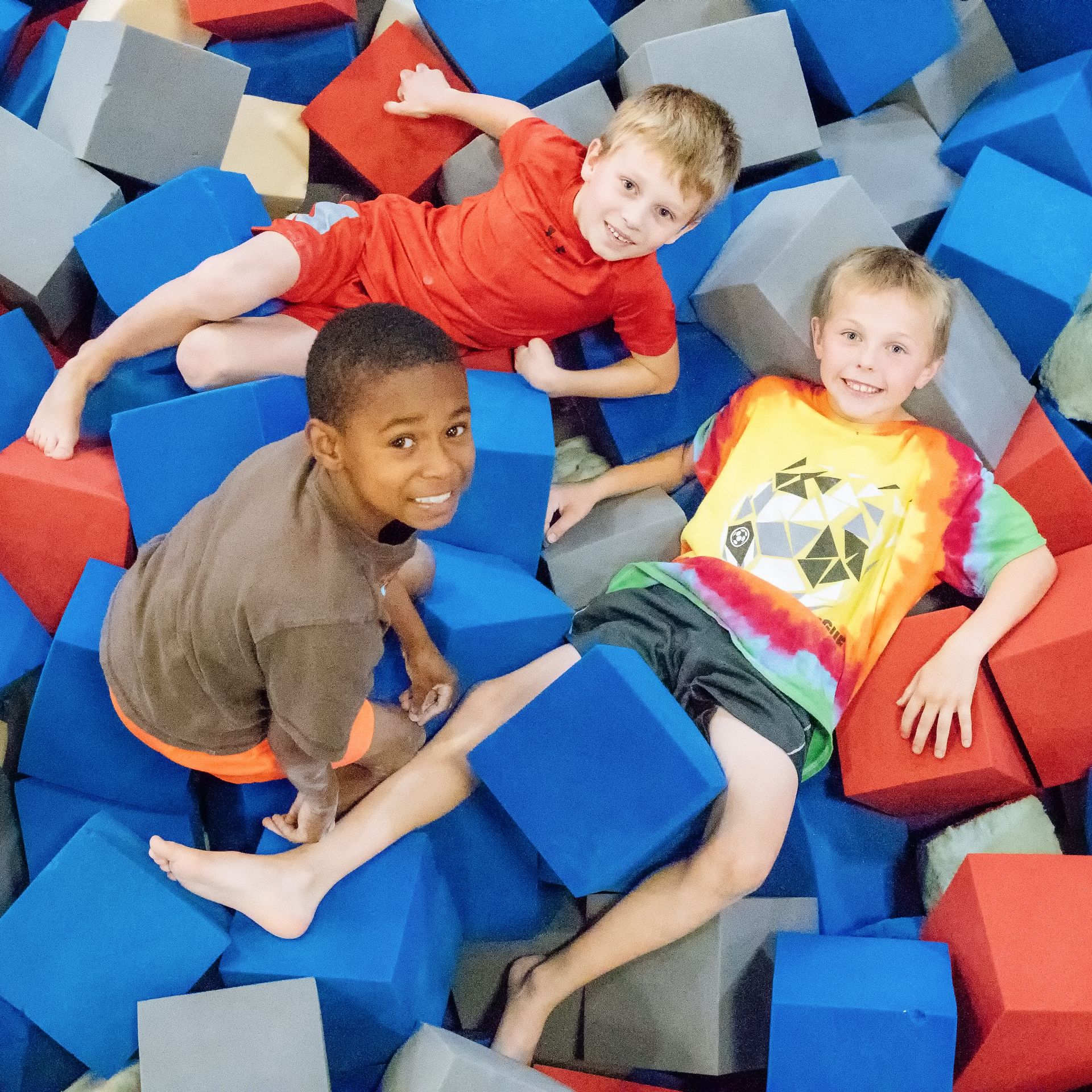 Three children lounging in a foam pit with red, blue, and gray blocks, smiling at the camera