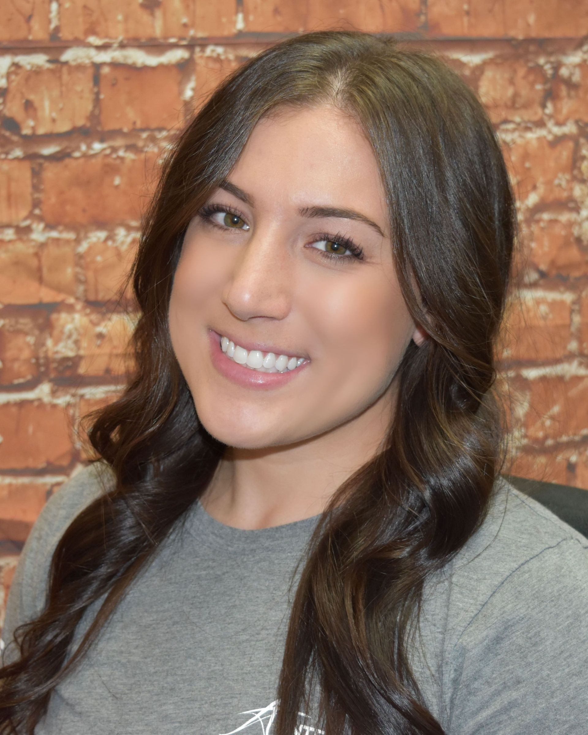 Smiling woman with long dark hair in a gray shirt standing before a brick wall