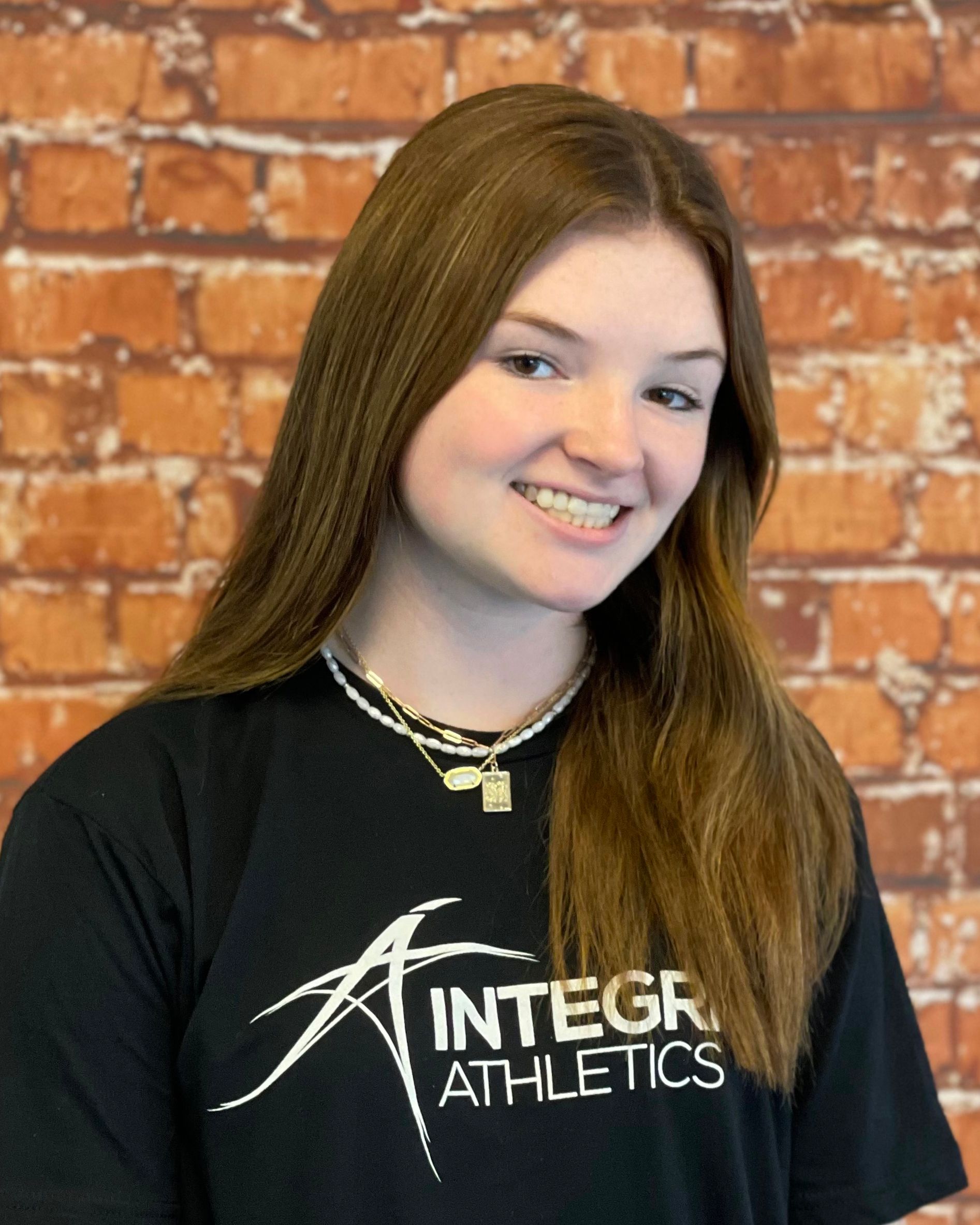 Smiling person in glasses wearing a dark T-shirt with a logo, standing against a brick wall.