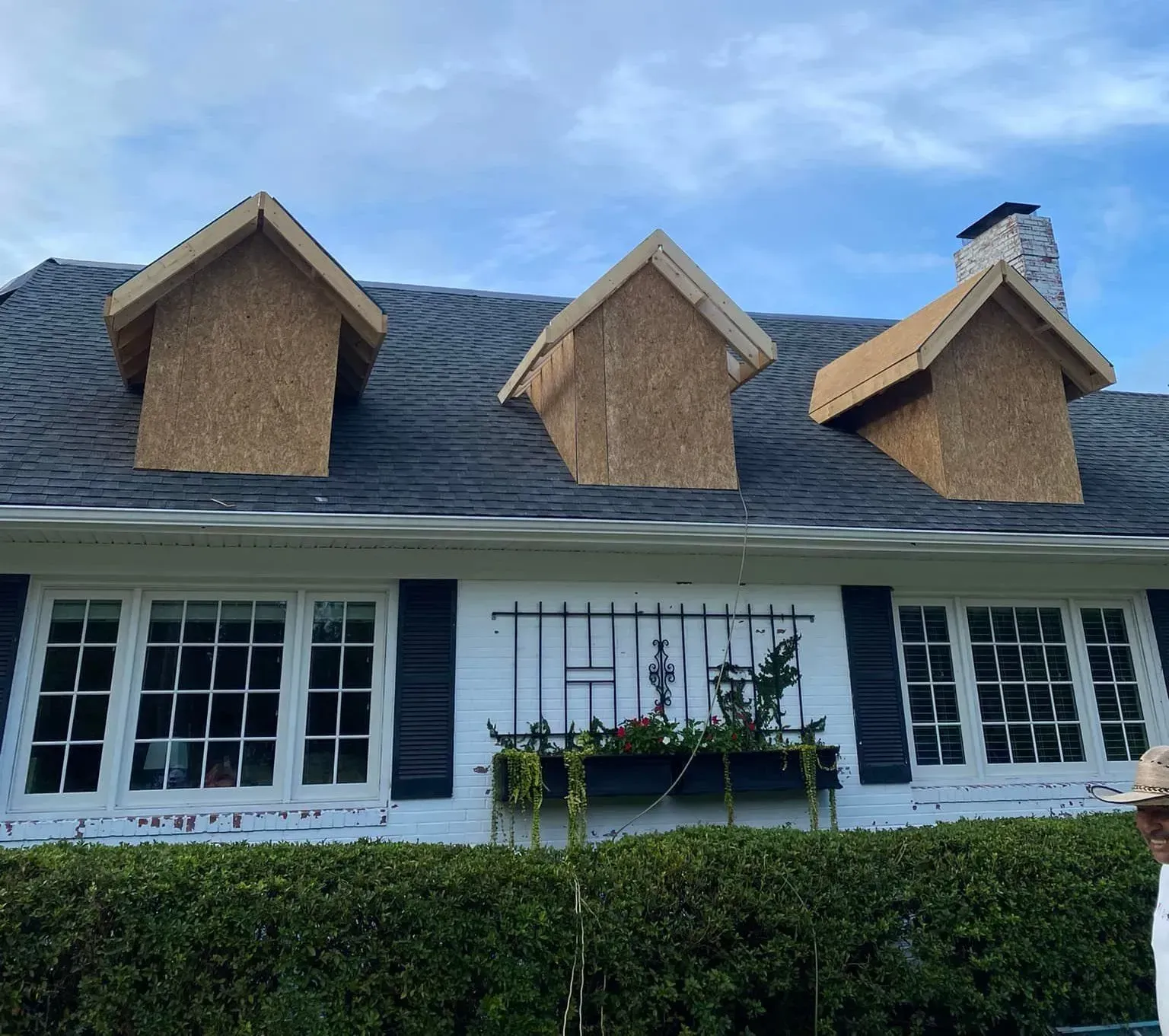 A white house with black shutters, three dormers under construction, and a green hedge.