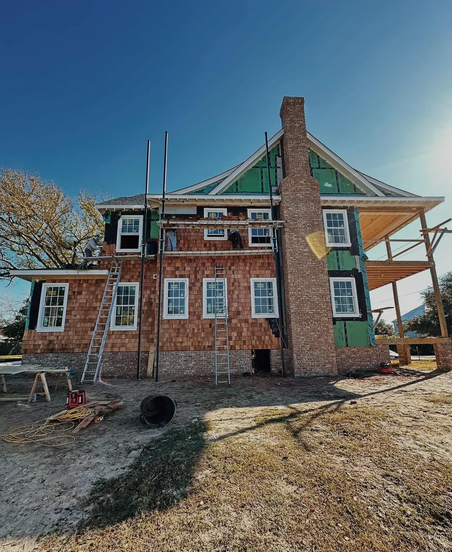 House under construction, brown siding, brick chimney, scaffolding, blue sky.