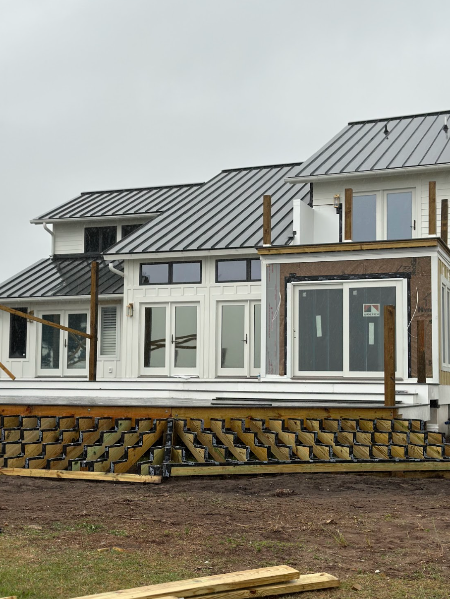 New home under construction, white siding, dark roof.  Wooden deck being built with stacked, zigzag pattern support.