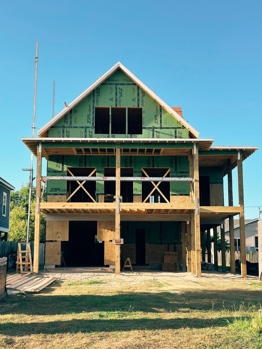 Two-story house under construction, wooden frame, green sheathing, blue sky.