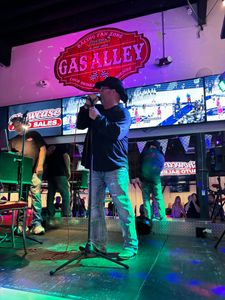 A man is singing into a microphone on a stage in front of a gas alley sign.
