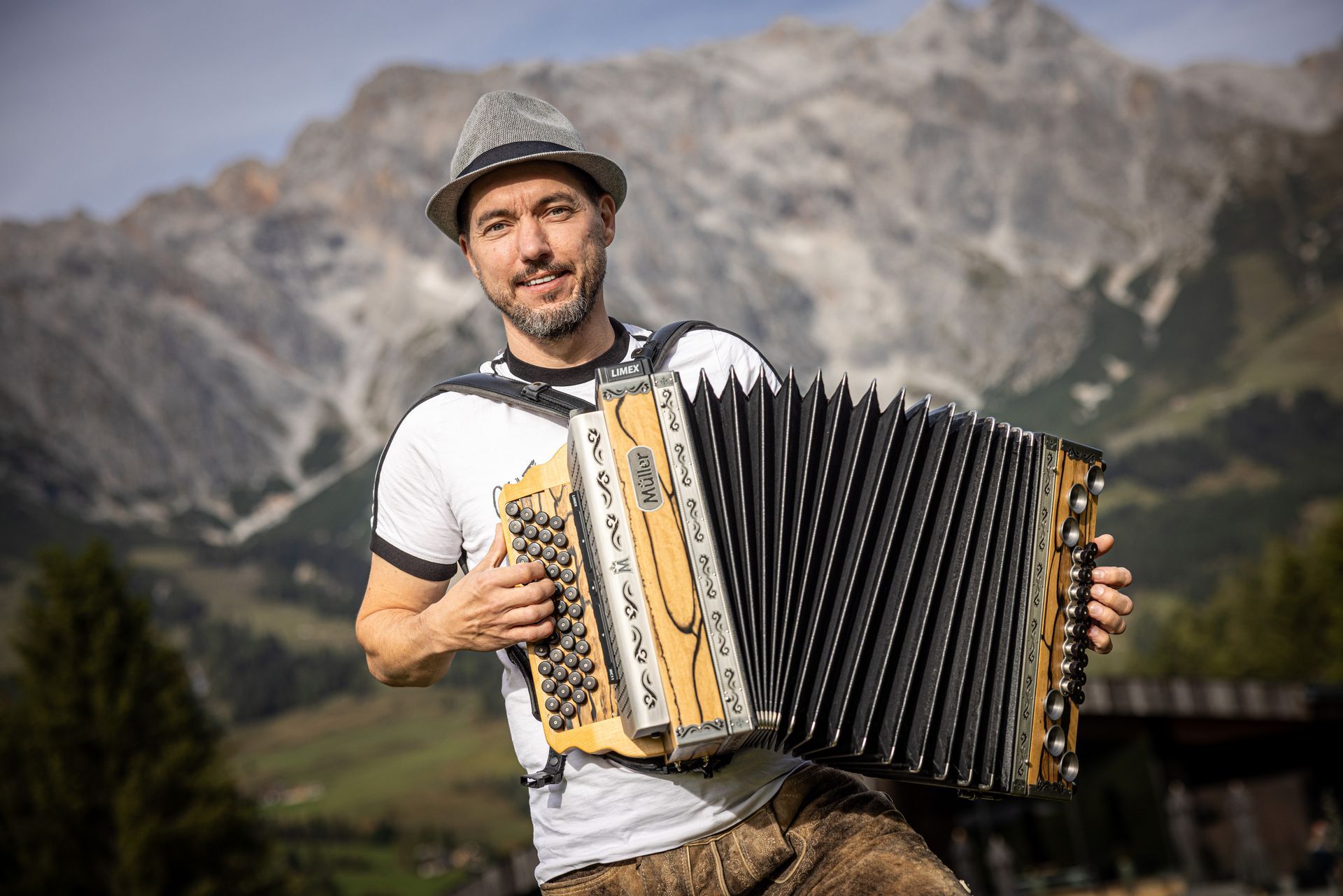Ein Mann spielt im Freien vor einer Bergkulisse Akkordeon, lächelt, trägt einen Hut und ein weißes Hemd.