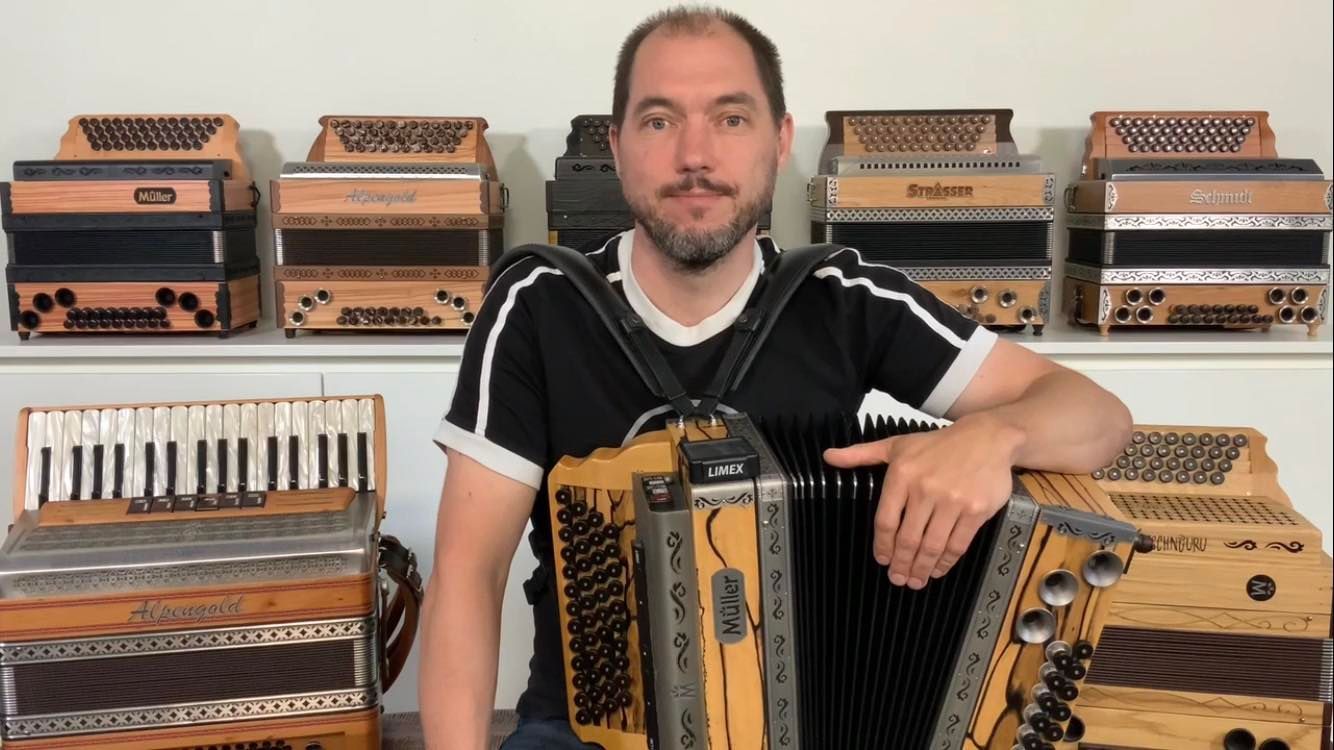 Ein Mann sitzt inmitten mehrerer Harmonikas; hinter ihm sind weitere Instrumente in Regalen ausgestellt.