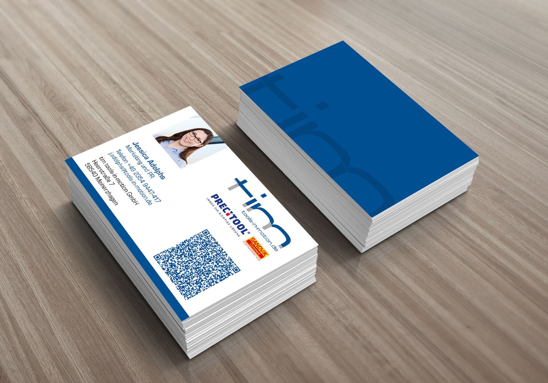 Zwei Stapel Visitenkarten auf einem Holztisch, einer davon mit blau-weißem Design und einem QR-Code. Anderer Blau mit Lack.