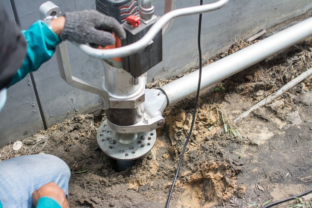 Worker Installing a Metal Pipe Fitting in Muddy Ground Beside a Building — Ground Screws SEQ Pty Ltd In Meridan Plains, QLD