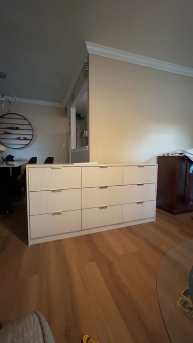 White dresser with many drawers in a living room, against a beige wall and wood floor.