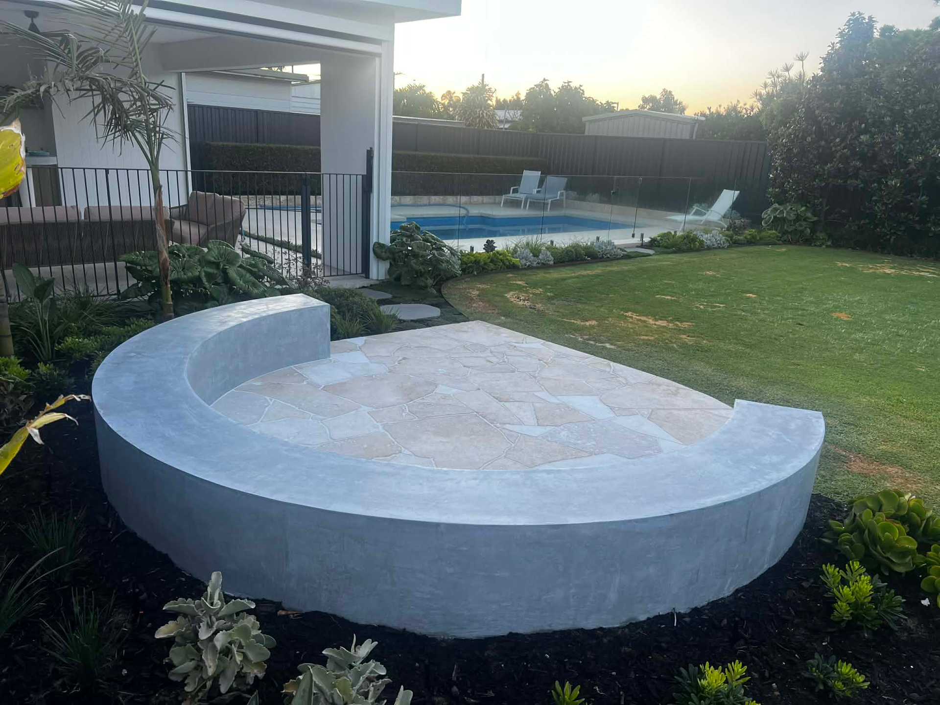 Rounded architectural concrete bench seat surrounding fire put area in landscaped backyard with grass area and a pool.