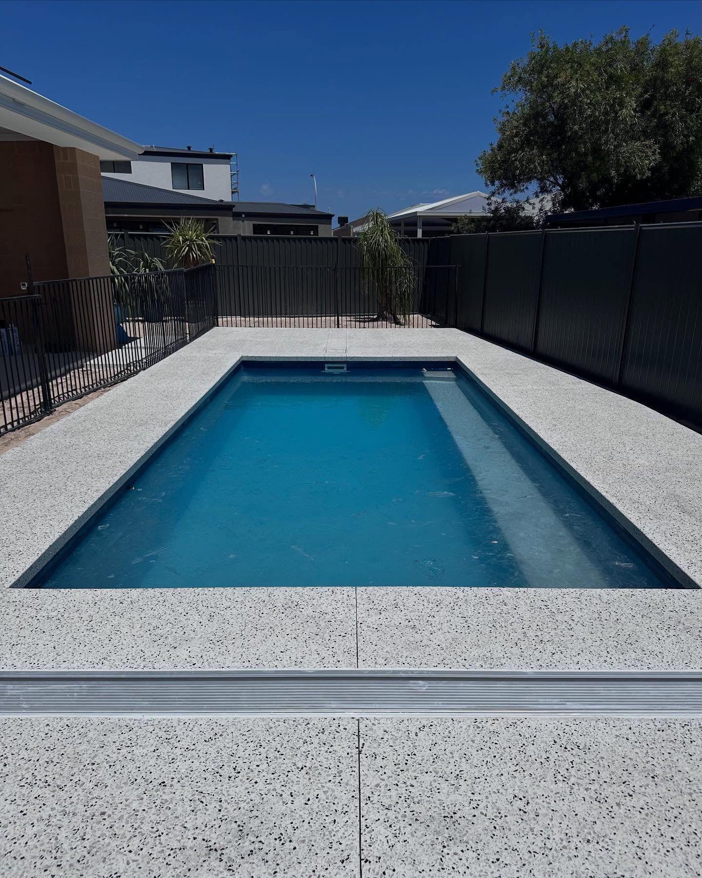 A rectangular backyard swimming pool with blue water and light gray speckled concrete surrounds
