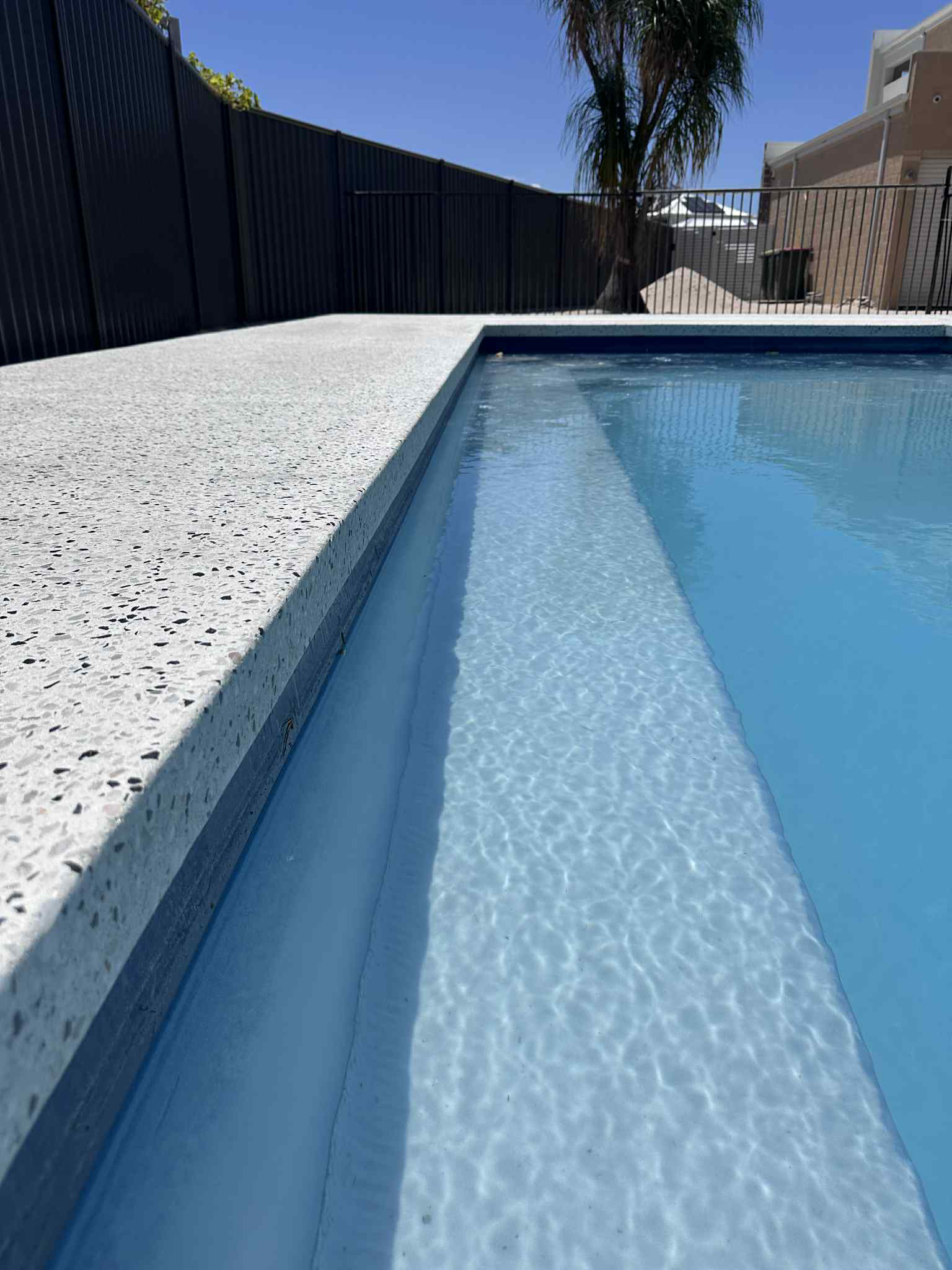 A side-view close-up of a swimming pool edge with speckled white honed concrete and clear blue water.