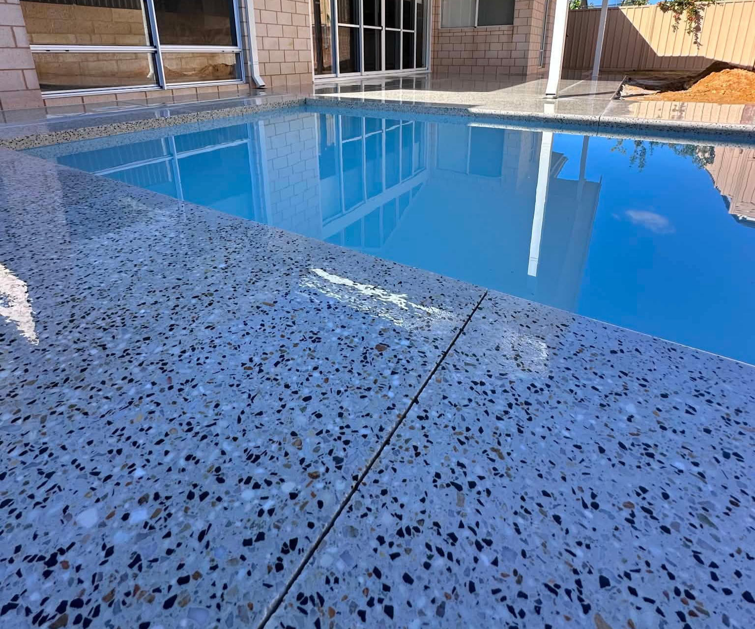 A backyard swimming pool with blue water and light-colored, honed concrete pool surrounding deck.