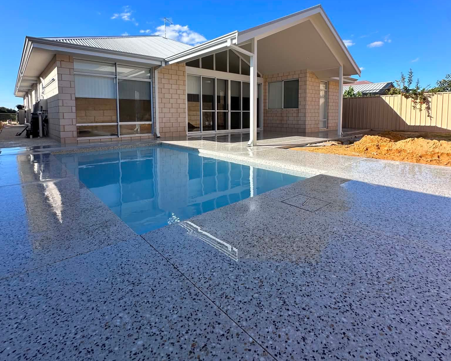 A house with a light-colored honed concrete, a covered patio, and a rectangular swimming pool with speckled concrete deck.