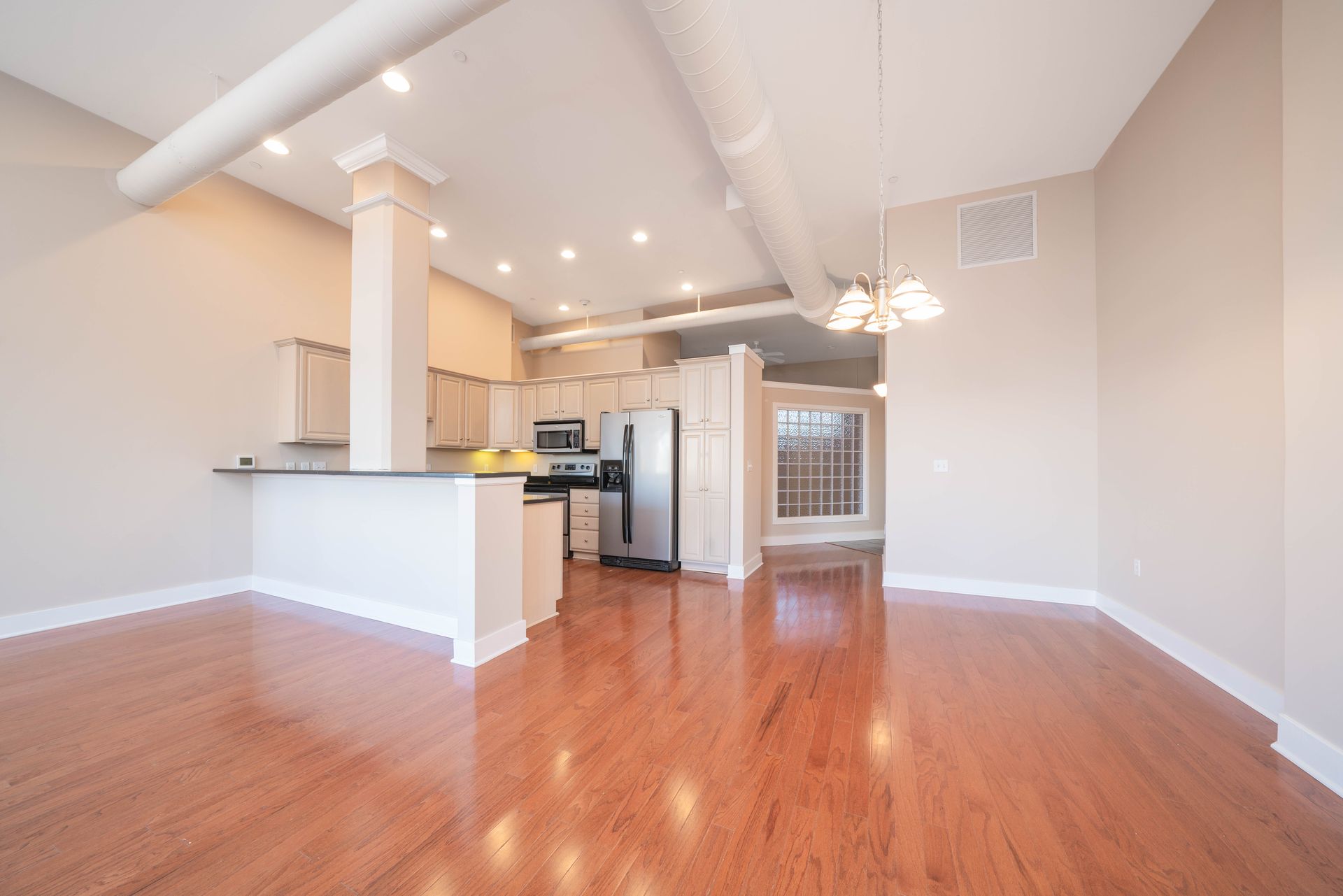 An empty room with hardwood floors and a kitchen in the background