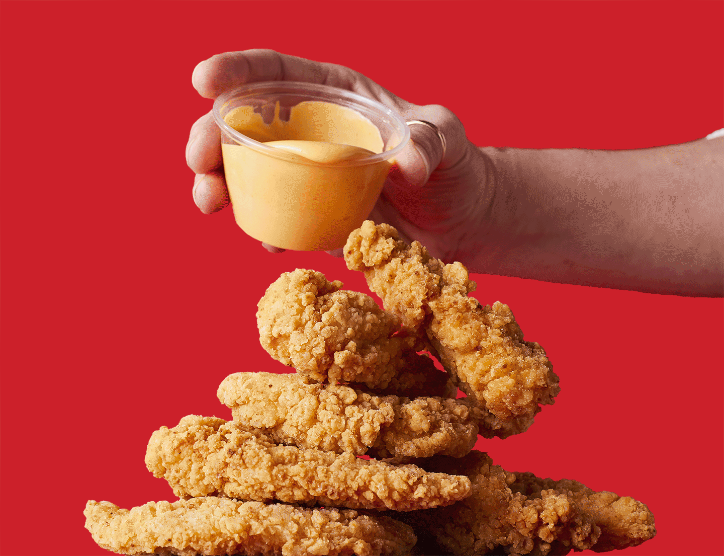 A person is holding a cup of sauce over a pile of chicken tenders.