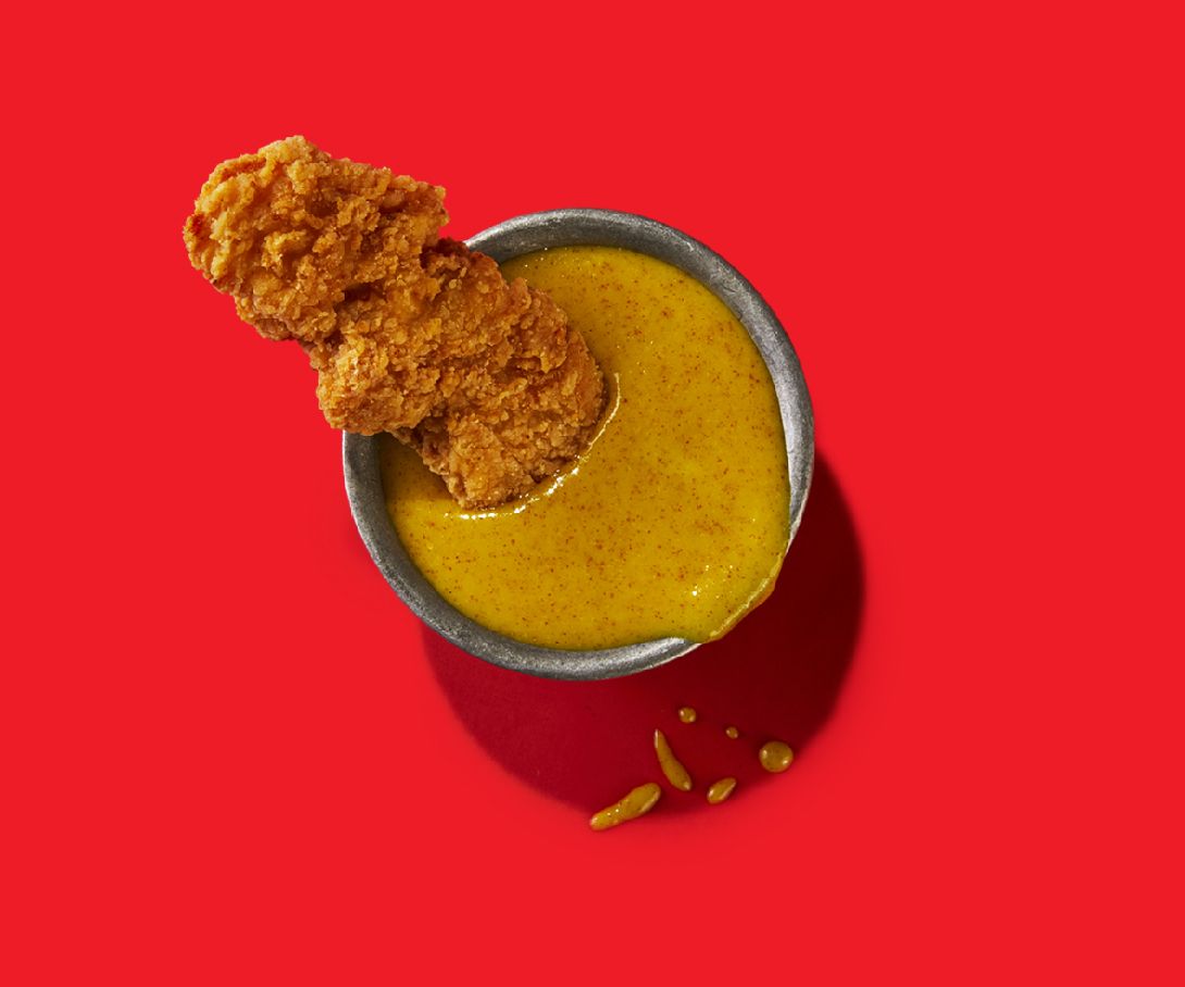 A chicken nugget is sticking out of a bowl of sauce on a red background.