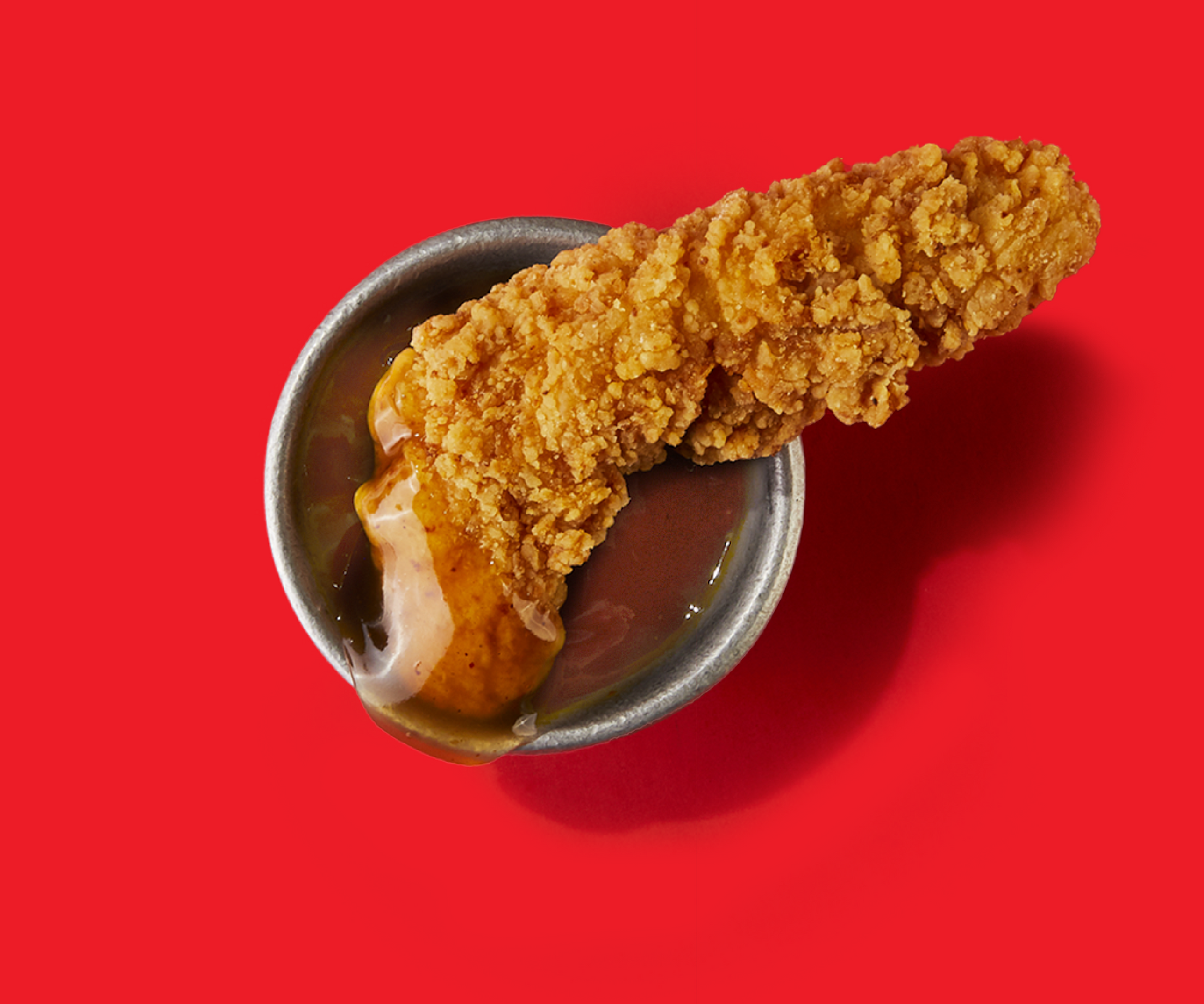 A piece of fried chicken is in a bowl of sauce on a red background.