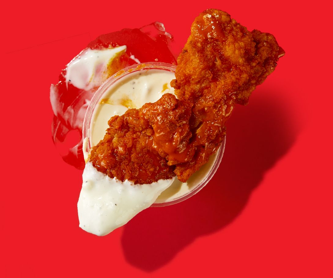 A piece of fried chicken is in a cup of sauce on a red background.