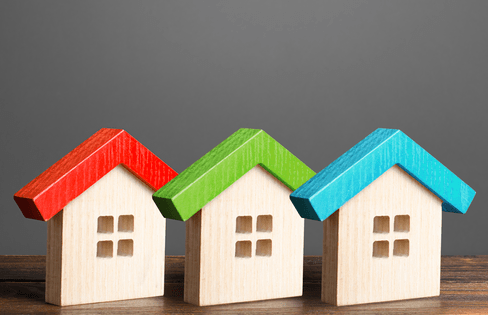 Three small wooden houses with red, green, and blue roofs on a wooden surface against a gray backdrop.