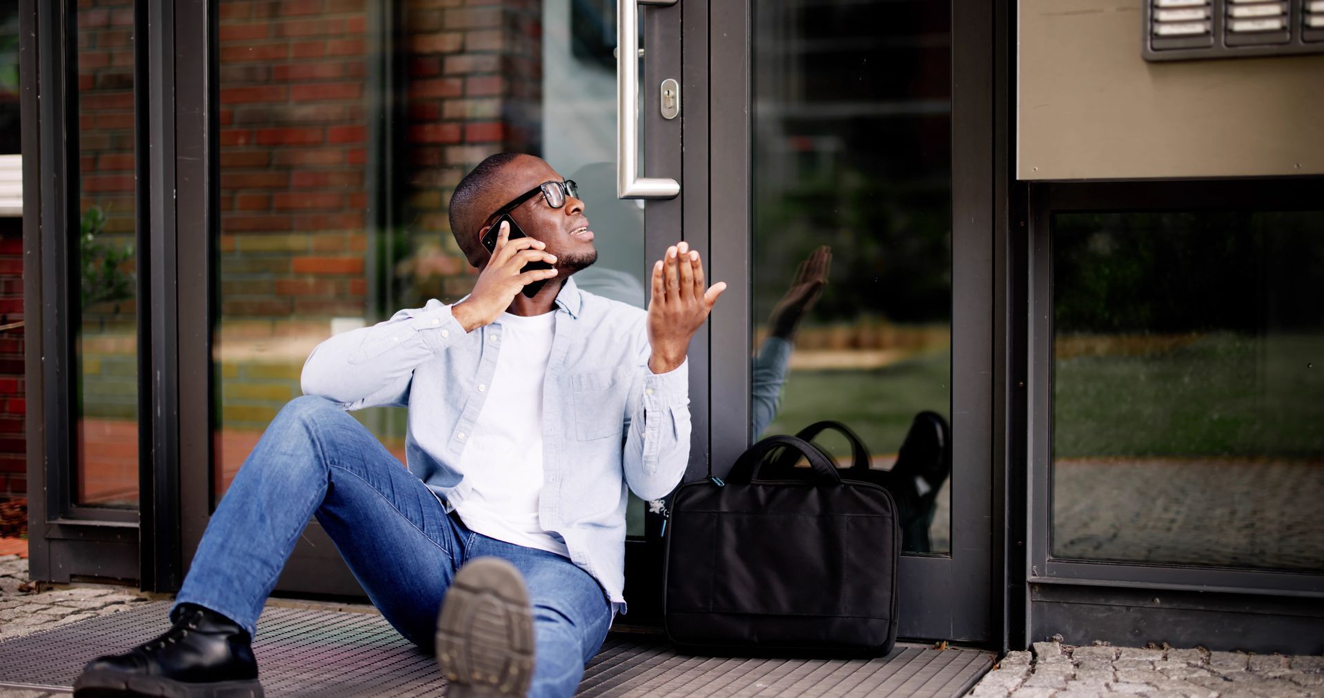 Man sits outside a building on the phone, looking upward, black bag beside him.