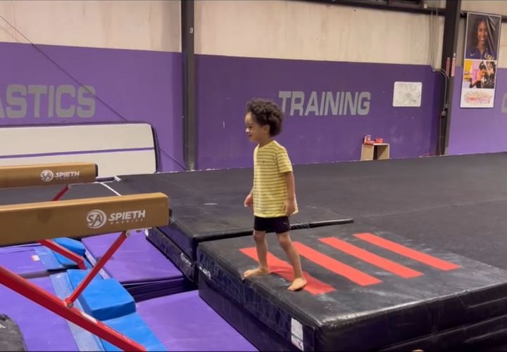 A young child in a yellow shirt and black shorts stands barefoot on a mat in a gymnastics gym.