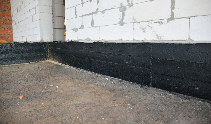 Black waterproofing material on a building's foundation, next to a concrete block wall and ground.