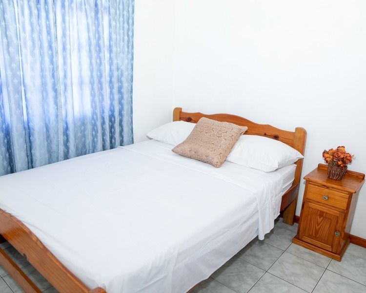 Small bedroom with a neatly made white bed, blue curtains, and a wooden nightstand with flowers.
