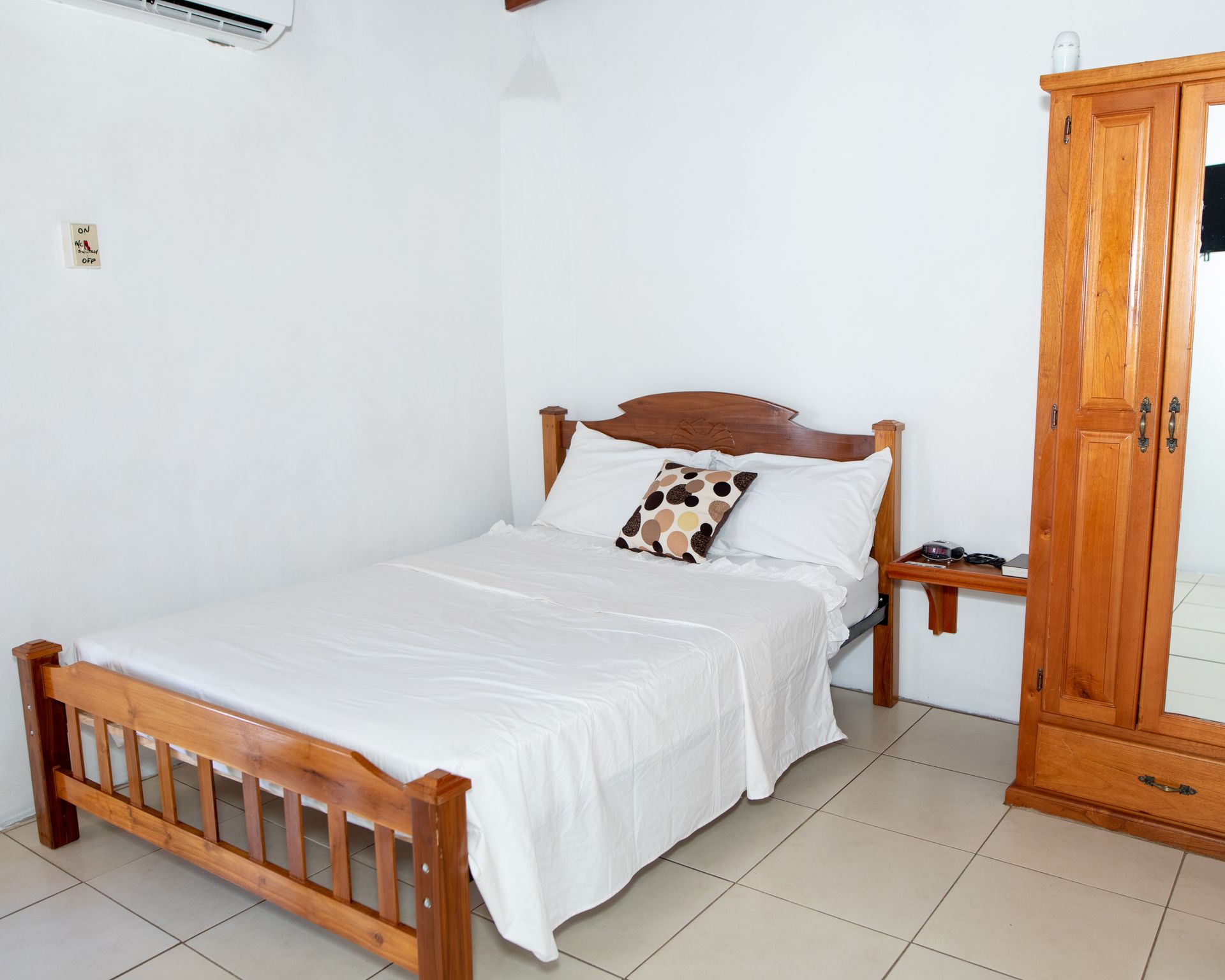 Simple bedroom with a wooden bed, white bedding, and a wardrobe on tiled floor.