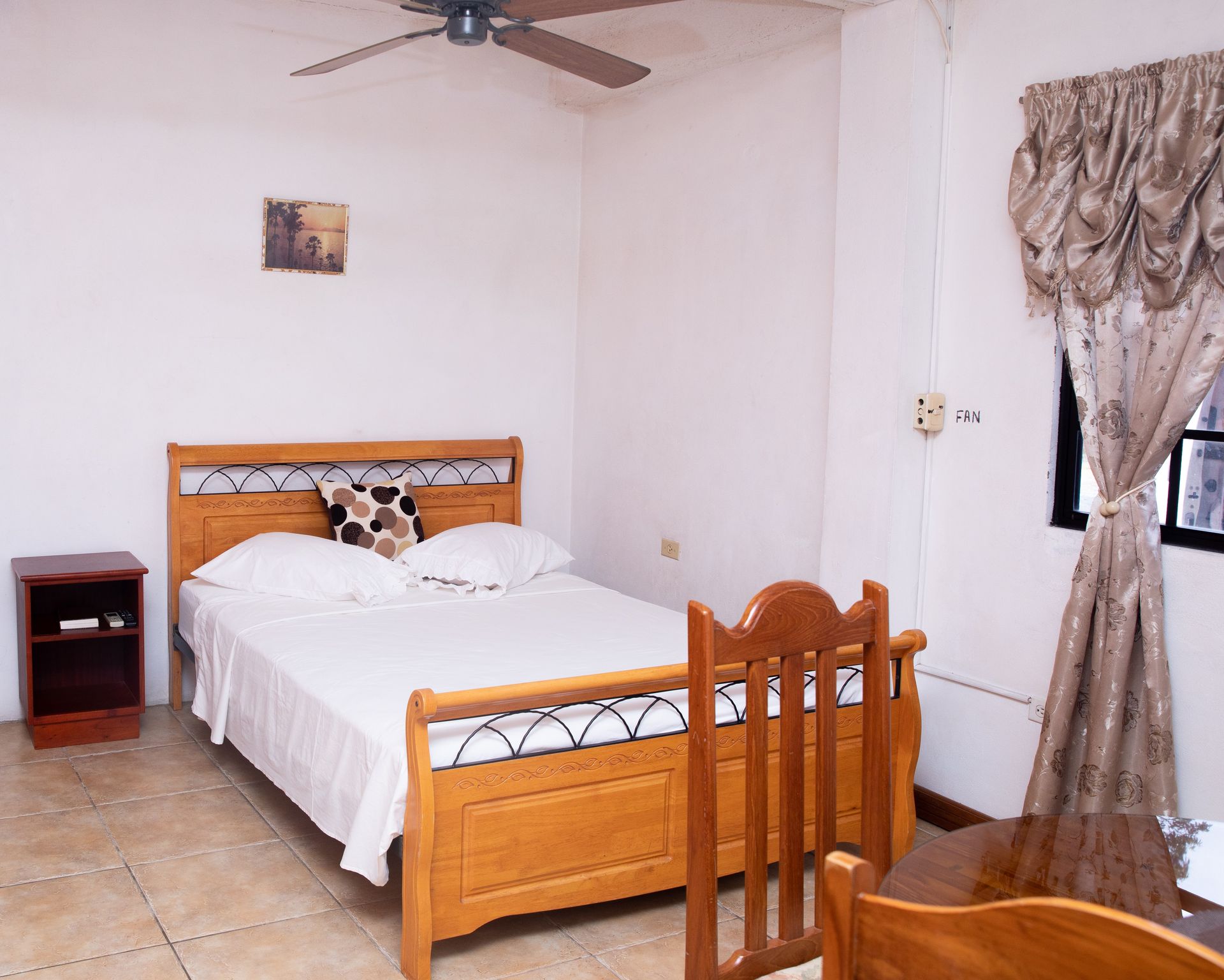 Small bedroom with wooden bed, ceiling fan, tiled floor, and curtained window.