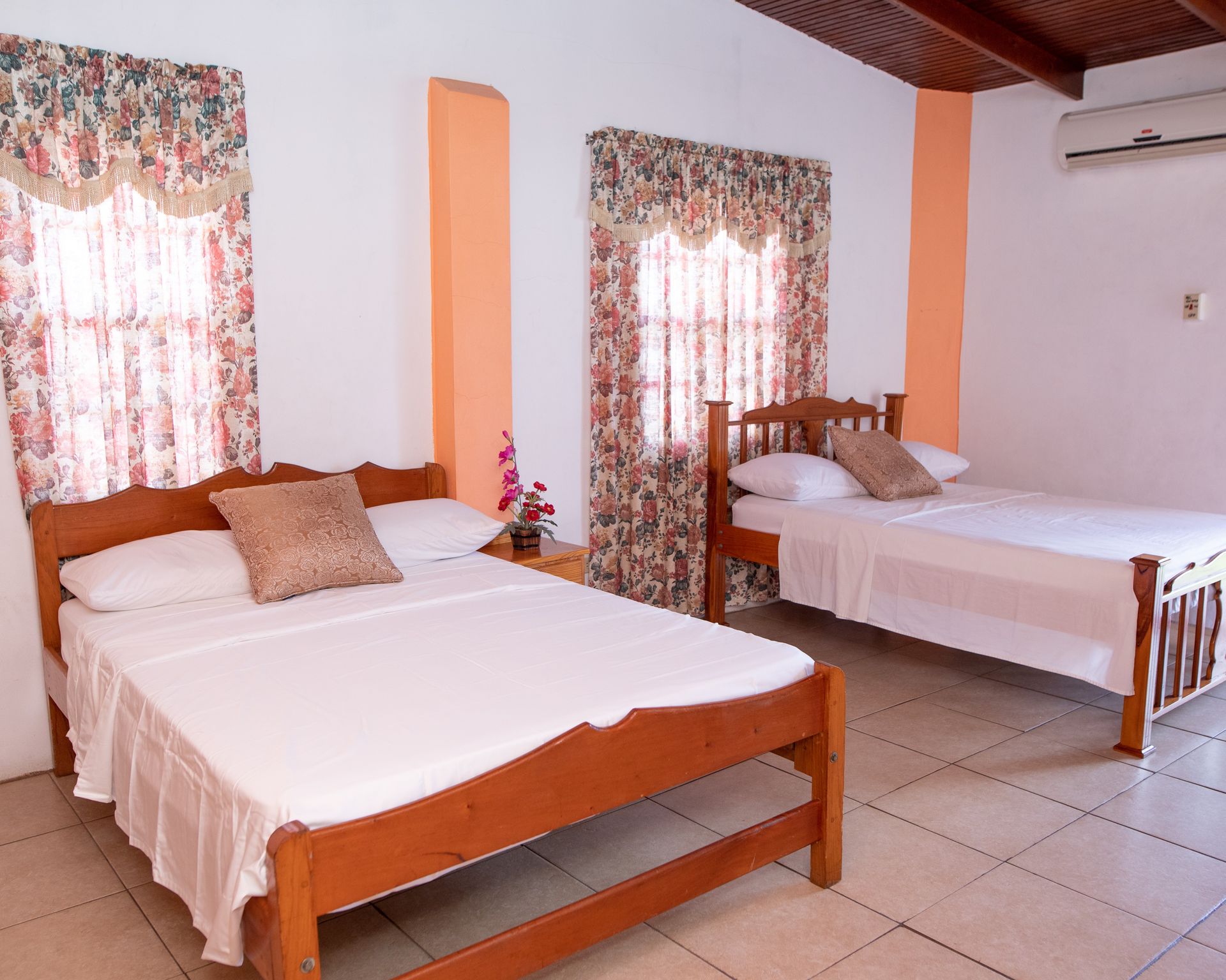 Small guest room with two wooden beds, white bedding, floral curtains, and tiled floor.