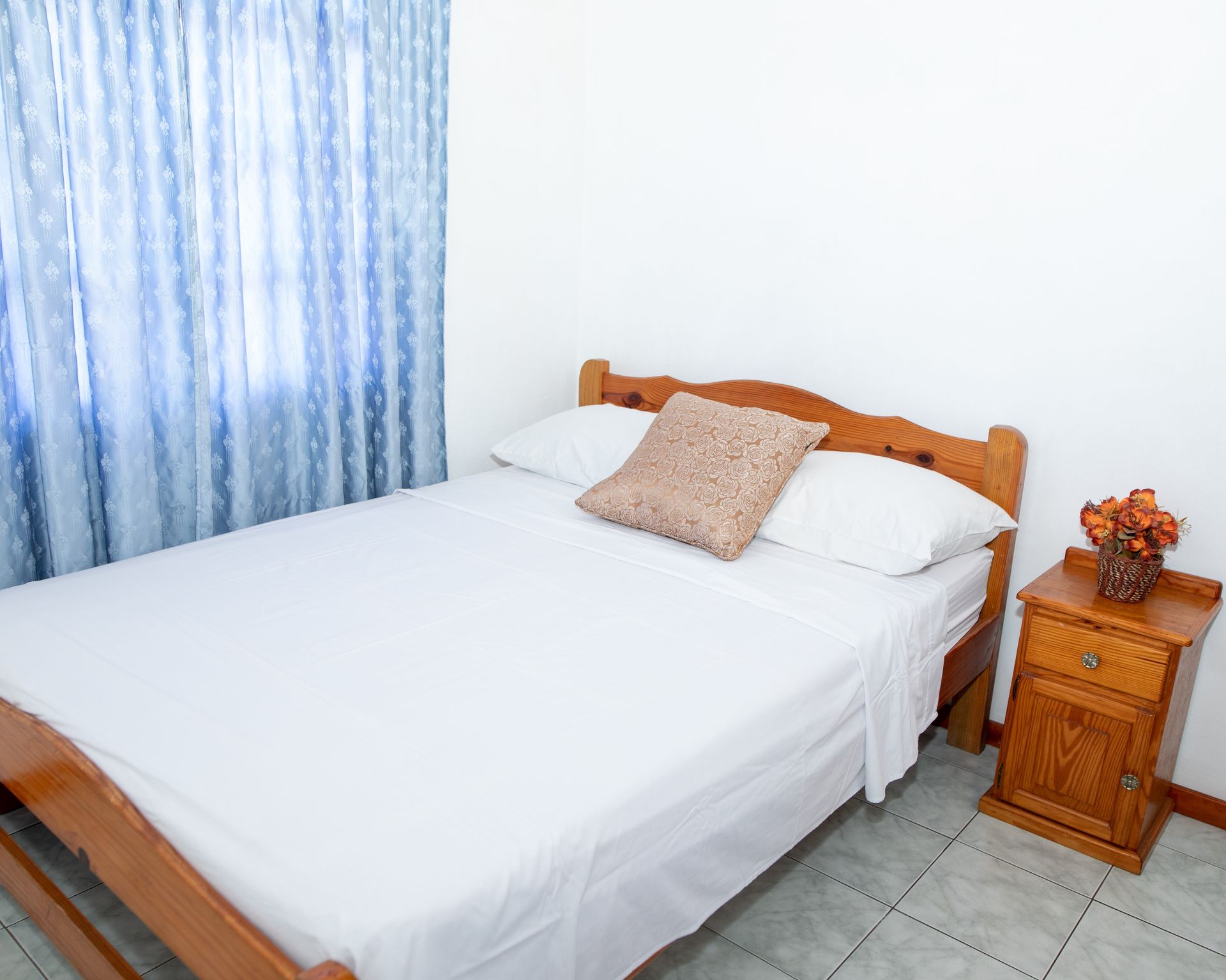 Small bedroom with a wooden bed, white bedding, blue curtains, and a nightstand with flowers.