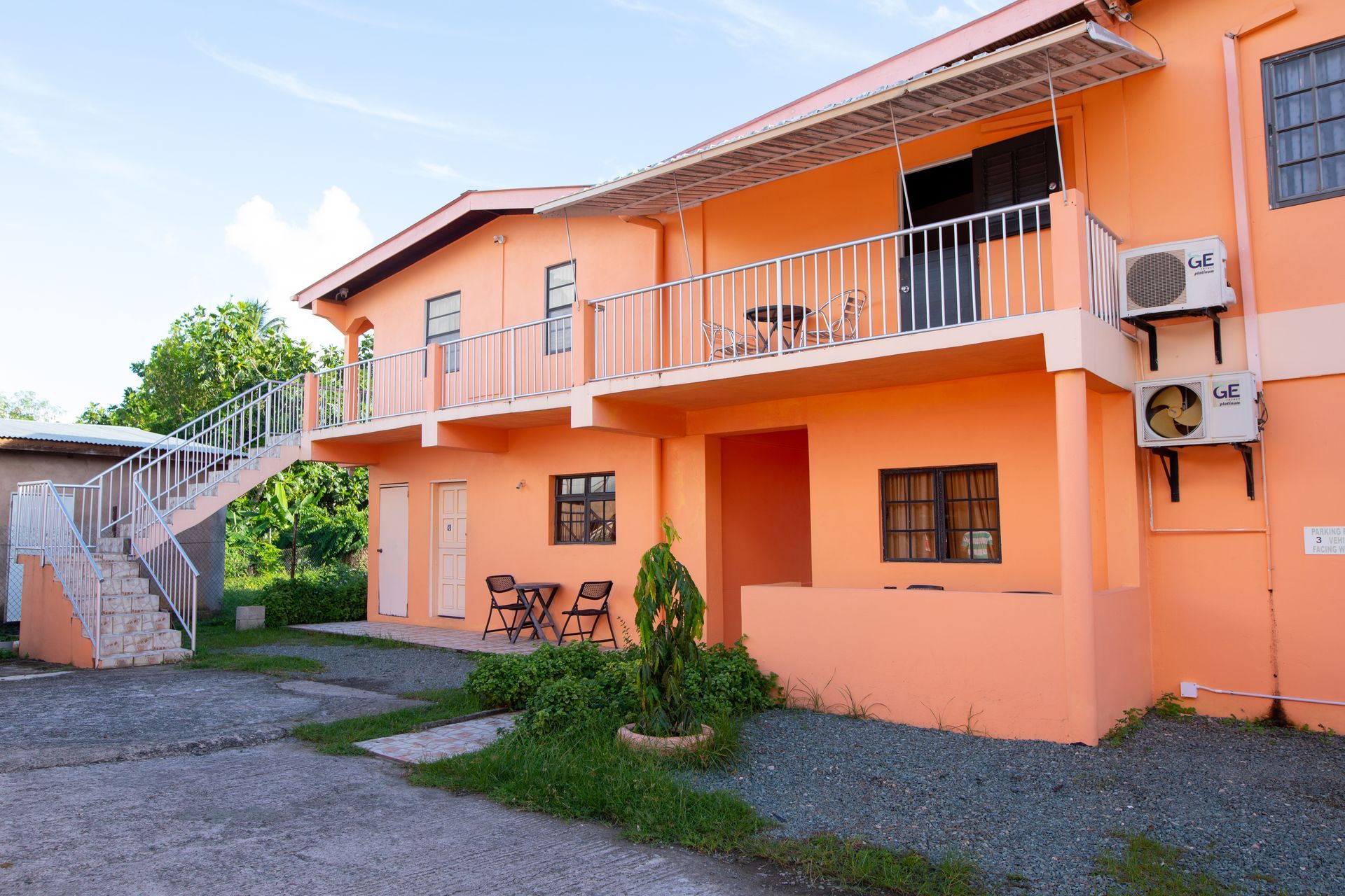 Orange apartment building with balconies, stairs, and parked air-conditioning units beside a gravel courtyard