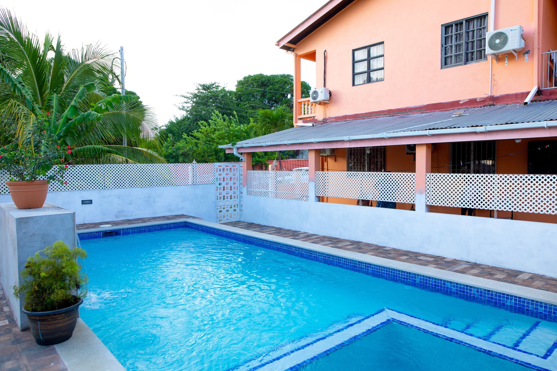 Backyard pool beside a peach house with blue water, tiled deck, and tropical greenery