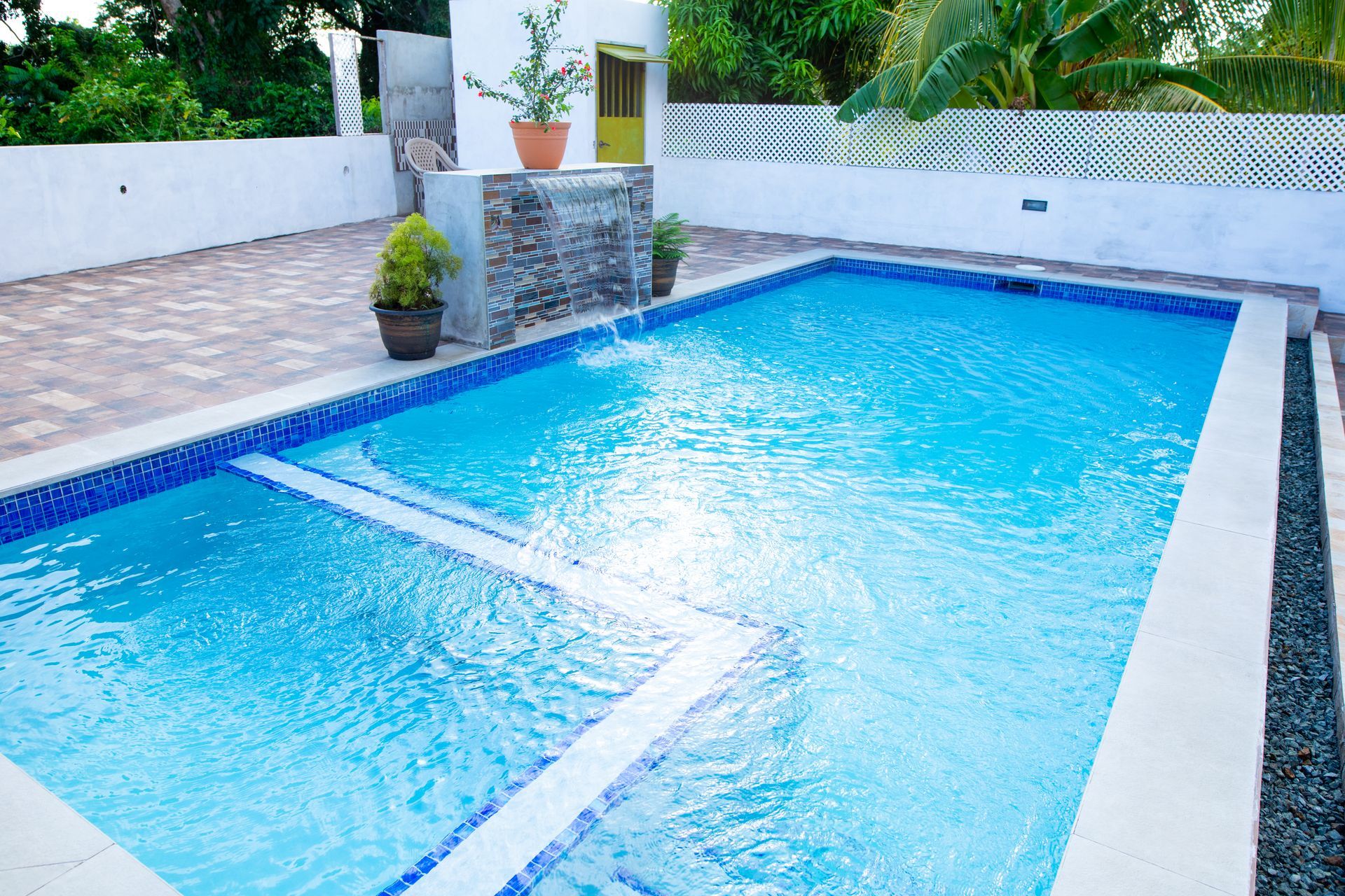 Bright blue swimming pool beside a tiled patio and white walls, surrounded by lush greenery.