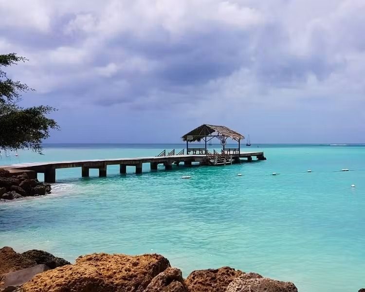 Turquoise sea with a wooden pier leading to a thatched hut under cloudy sky