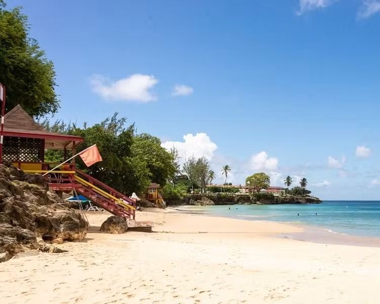 Sunny tropical beach with white sand, turquoise water, and a small red-roofed hut by trees