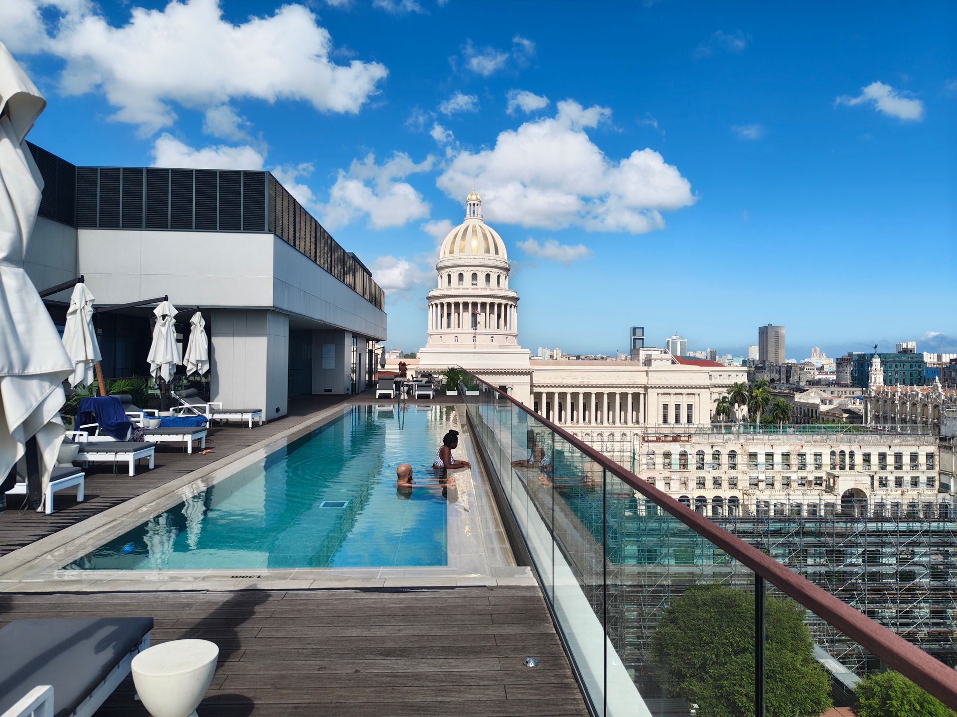 Rooftop infinity pool with loungers overlooking a domed city building and skyline under a blue sky