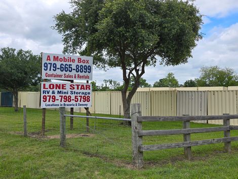 Two business signs in a grassy yard beside a fence and tree, with a wooden gate in front.