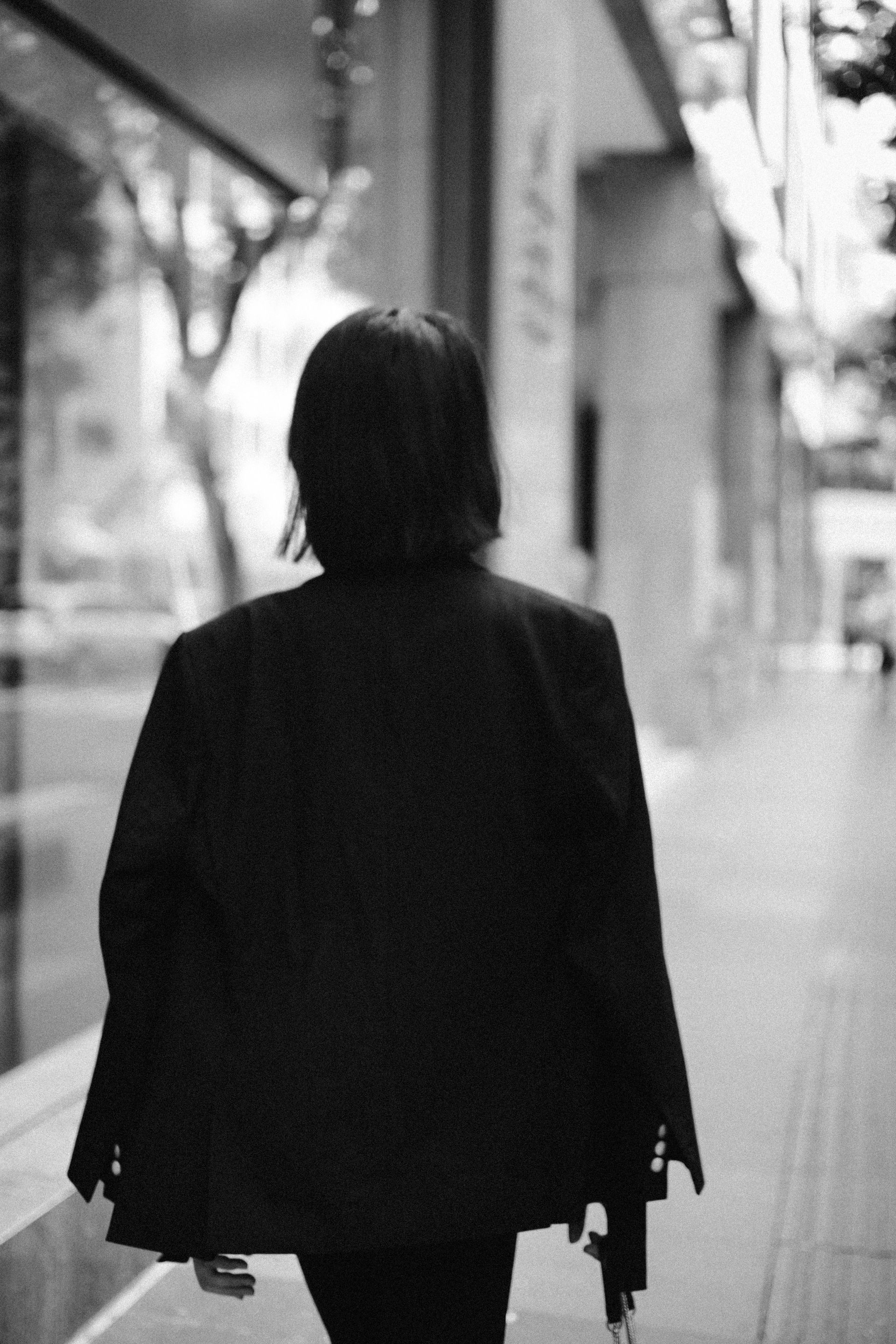 Person walking away on a city sidewalk beside storefronts, wearing a dark coat.