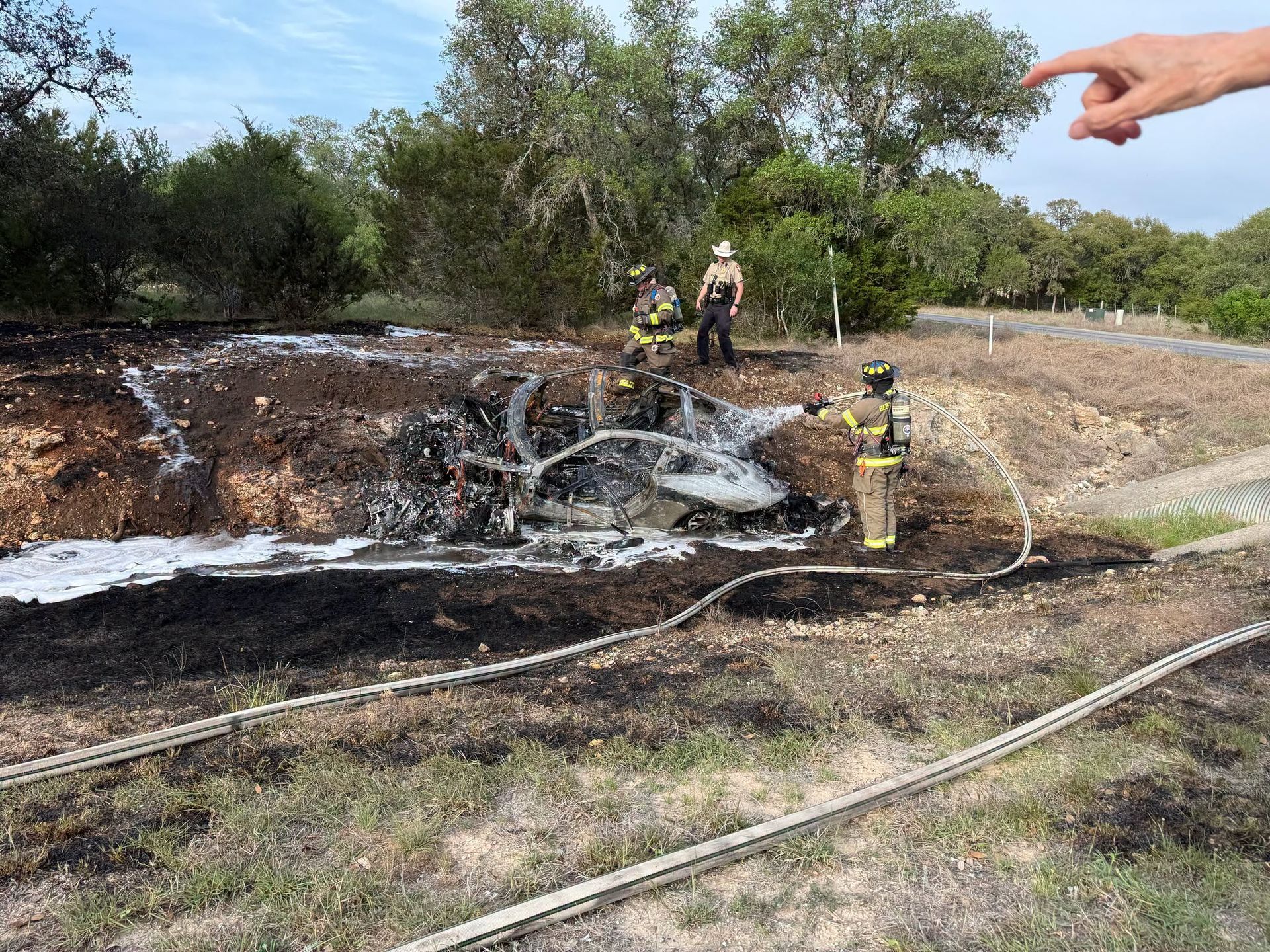 Firefighters spraying water on a burned field near a smoking brush fire, with a pointing hand in the foreground