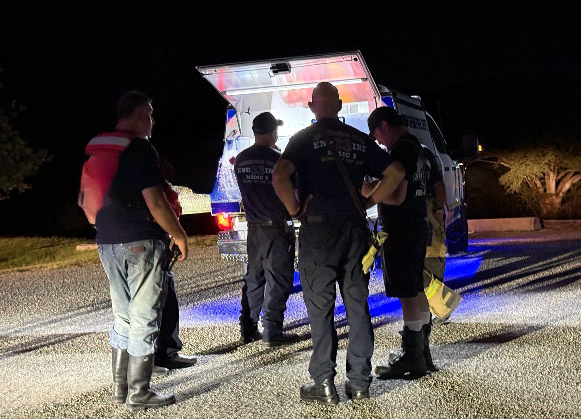 Fire fighters standing beside an open vehicle at night on a roadside with blue emergency lights.