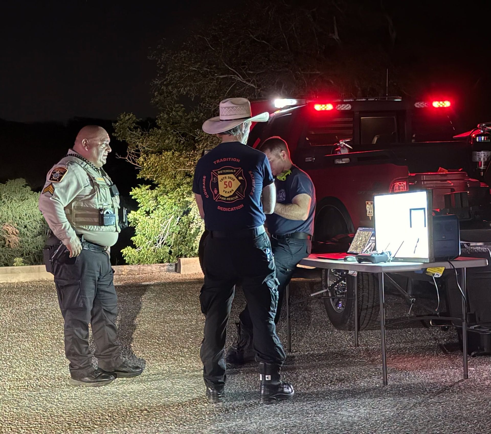 Three people stand by a lit table near a vehicle at night, with flashing emergency lights behind them.