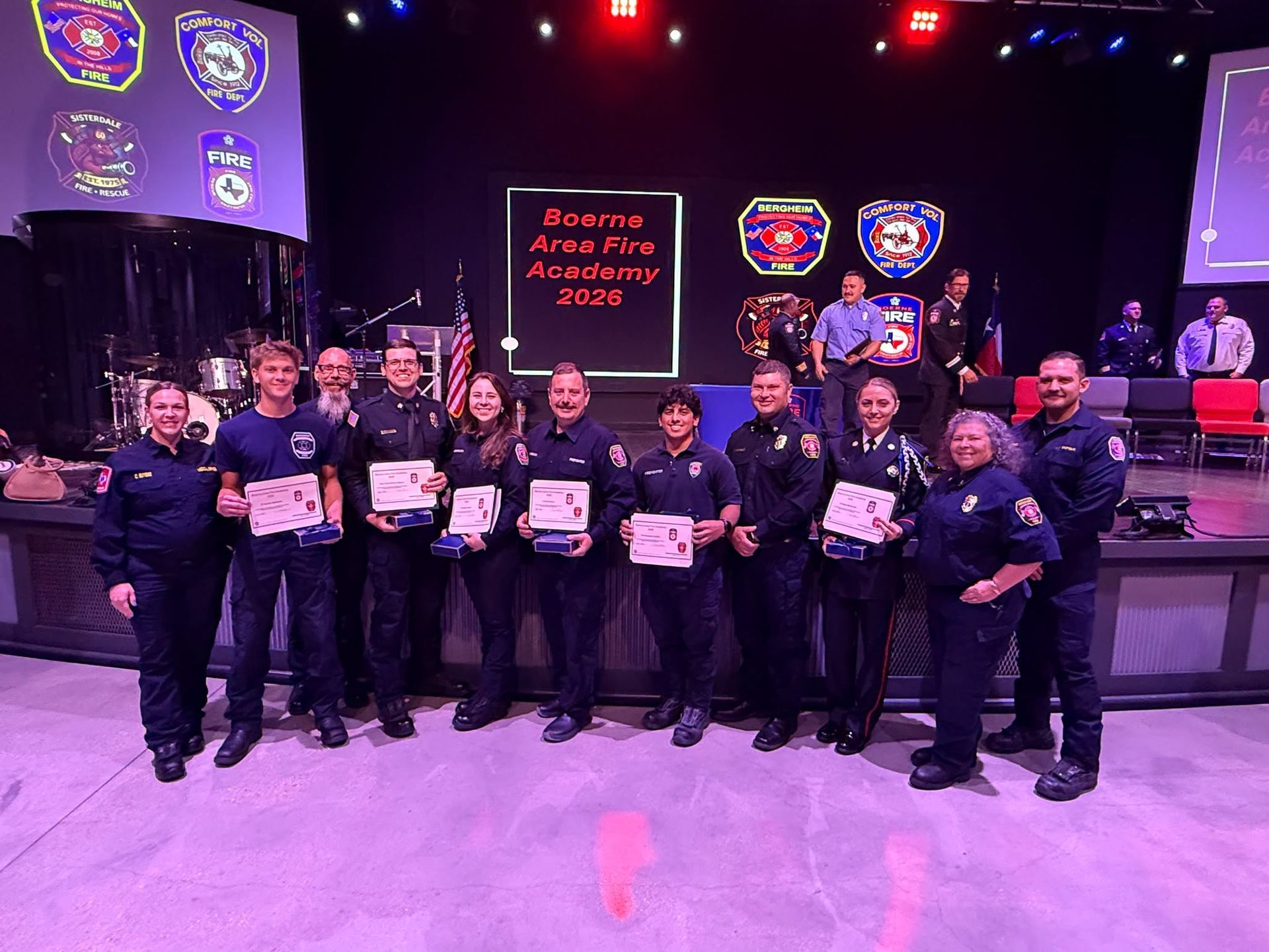 Group of uniformed award recipients holding certificates on a stage with police-style banners and lights