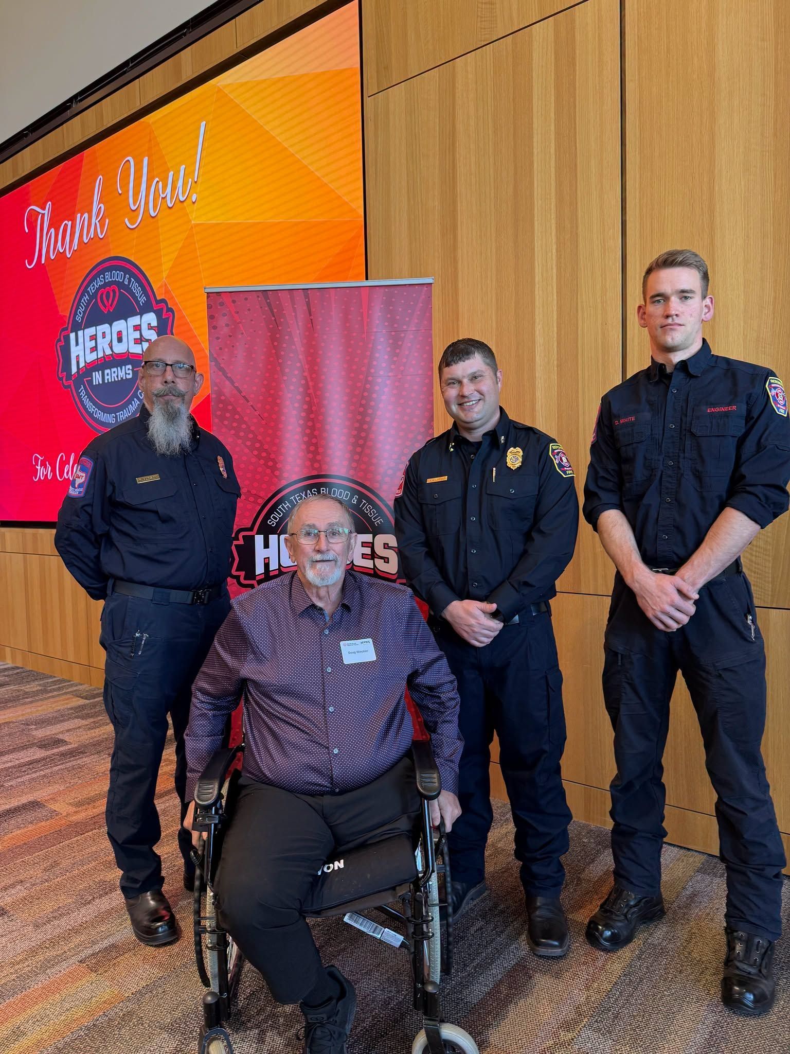 Four people in a wood-paneled room, one seated in a wheelchair, with a red banner and award sign behind them.