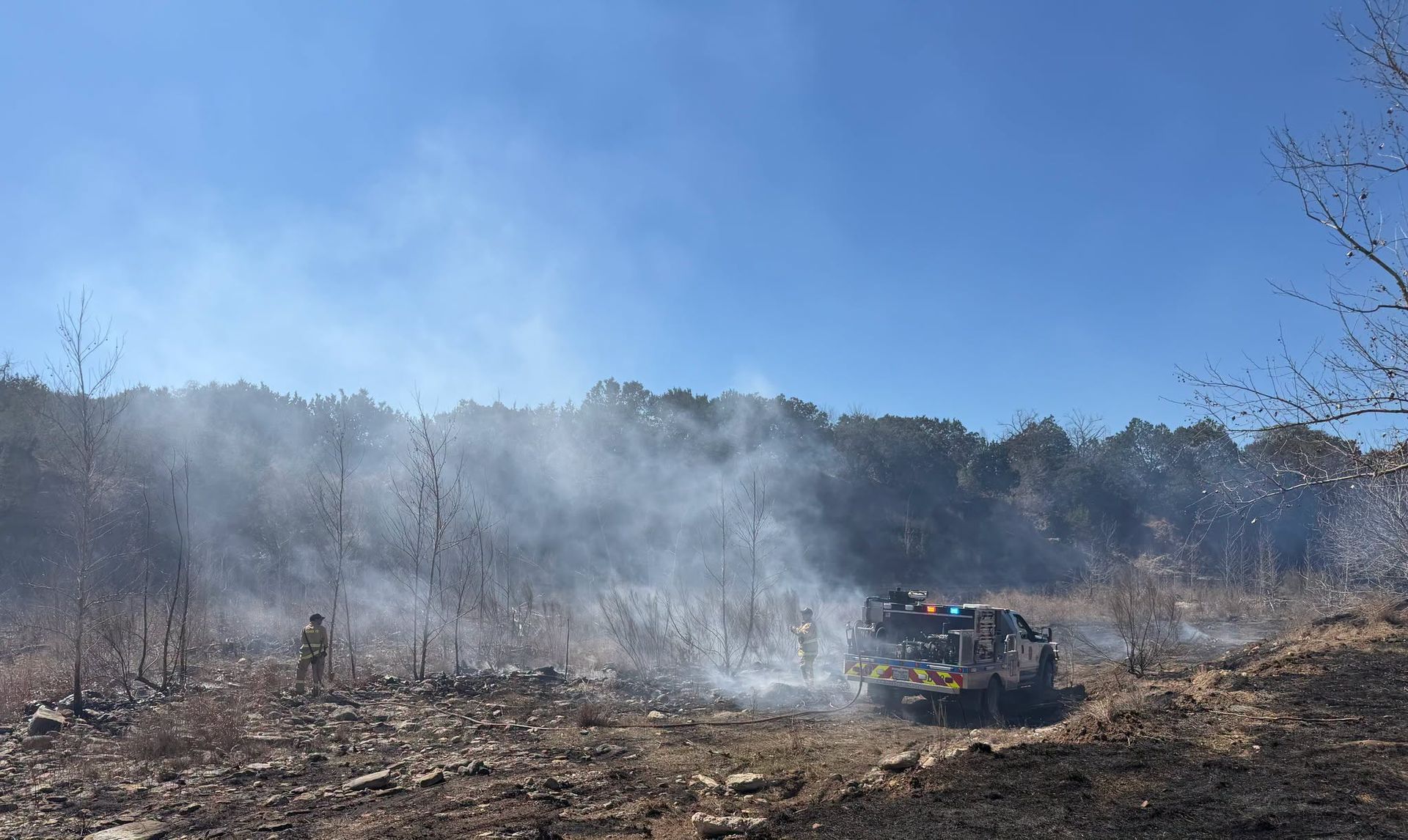 Smoke rising from a burned field under a blue sky, with a vehicle near the center.