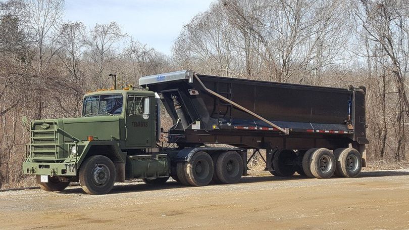 Green dump truck with raised black trailer parked on a dirt road in a wooded area